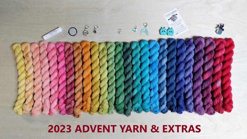 May include: A collection of twenty-four mini yarn skeins arranged in a rainbow gradient, accompanied by small metal stitch markers, a lip balm, two teal whale-shaped stitch stoppers, and a small card labeled 2023 ADVENT YARN & EXTRAS.