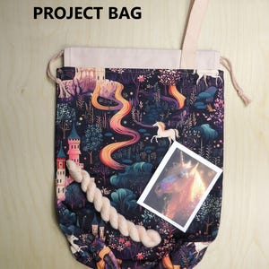 May include: A drawstring project bag featuring a dark fabric with a colorful print of unicorns, castles, and trees. The bag has a light-colored top section, a single strap, and includes a small skein of yarn and a rectangular card with a unicorn image. Text reads 2025 PROJECT BAG.