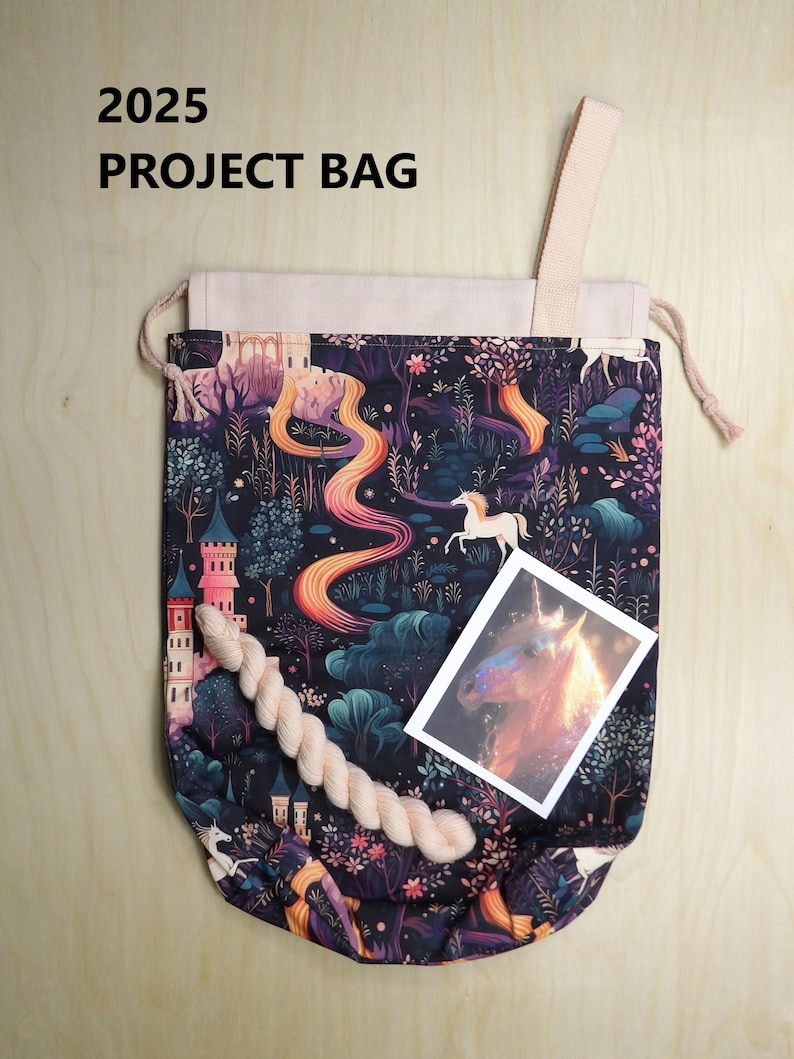 May include: A drawstring project bag featuring a dark fabric with a colorful print of unicorns, castles, and trees. The bag has a light-colored top section, a single strap, and includes a small skein of yarn and a rectangular card with a unicorn image. Text reads 2025 PROJECT BAG.