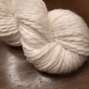 May include: A skein of white yarn, made of natural fibers, is shown in a close-up view. The yarn is soft and fluffy, and it is perfect for knitting or crocheting.