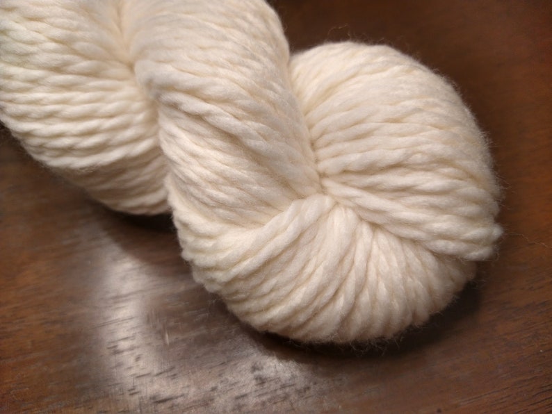 May include: A skein of white yarn, made of natural fibers, is shown in a close-up view. The yarn is soft and fluffy, and it is perfect for knitting or crocheting.