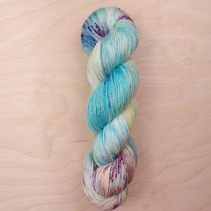May include: A skein of hand-dyed yarn in shades of turquoise, teal, purple, and cream. The yarn is twisted into a spiral shape, showcasing the colour variations. The yarn is likely made of wool or a similar fibre, perfect for knitting or crocheting projects.