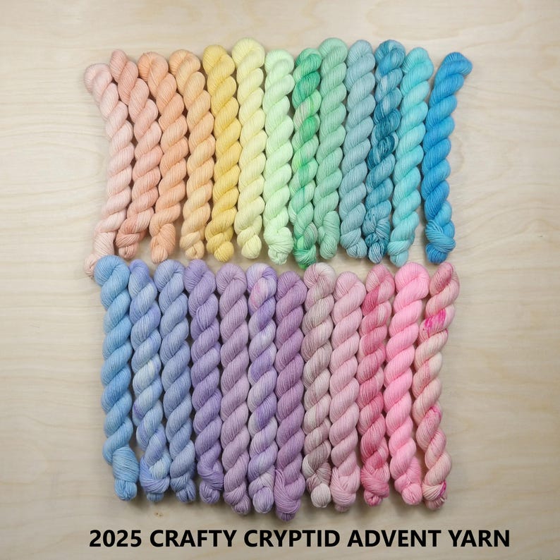 May include: A collection of twenty-four twisted mini skeins of yarn arranged in two rows, featuring a gradient of pastel colors including peach, yellow, green, blue, purple, and pink. The text 2025 CRAFTY CRYPTID ADVENT YARN is printed below.