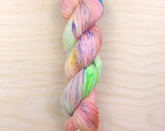 May include: A skein of hand-dyed yarn in a twisted, vertical arrangement. The yarn features a blend of pastel colors, including pink, blue, green, and yellow, with speckled accents. The yarn is displayed against a light wood grain background.