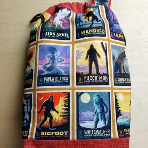 May include: A drawstring bag featuring a grid of colorful rectangular illustrations of cryptids and national parks, including Zion Angel, Wendigo, Pavla Blanca, Yucca Man, Bigfoot, and Whitewalker. The bag has a red textured fabric base and top trim.