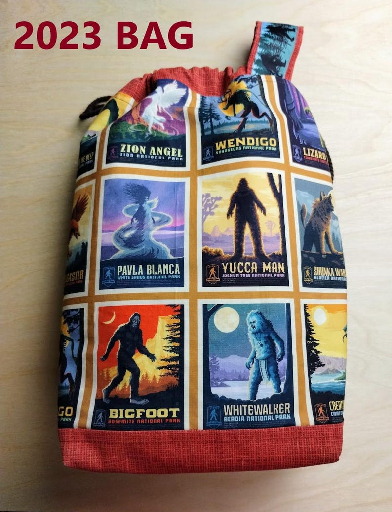 May include: A drawstring bag featuring a grid of colorful rectangular illustrations of cryptids and national parks, including Zion Angel, Wendigo, Pavla Blanca, Yucca Man, Bigfoot, and Whitewalker. The bag has a red textured fabric base and top trim.