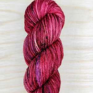 May include: A skein of deep red yarn with subtle purple and pink flecks. The yarn is twisted and piled on a light wood surface.