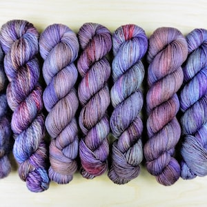 May include: A set of seven skeins of hand-dyed yarn in shades of purple, blue, and brown. The yarn is arranged in a row, with the skeins overlapping slightly.