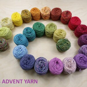 May include: A collection of twenty-four small yarn balls arranged in a spiral pattern, featuring a gradient of colors from yellow and orange to red, purple, blue, and green. The text 2024 ADVENT YARN is printed in purple at the bottom.