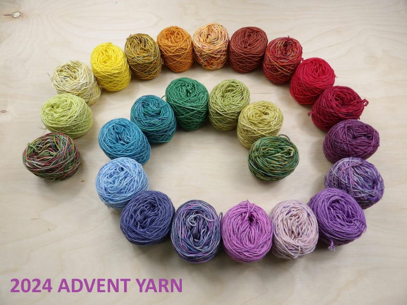 May include: A collection of twenty-four small yarn balls arranged in a spiral pattern, featuring a gradient of colors from yellow and orange to red, purple, blue, and green. The text 2024 ADVENT YARN is printed in purple at the bottom.