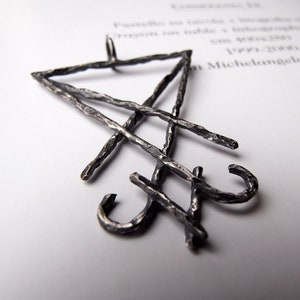 May include: A silver-colored pendant with a geometric design. The pendant features a triangle shape at the top, with intersecting lines and curved elements. The pendant is made of a textured metal and is displayed on a white surface.