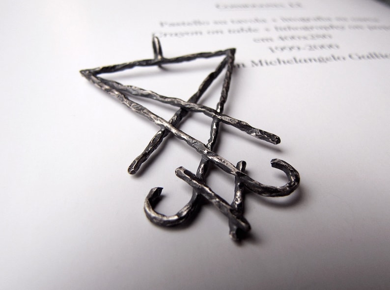 May include: A silver-colored pendant with a geometric design. The pendant features a triangle shape at the top, with intersecting lines and curved elements. The pendant is made of a textured metal and is displayed on a white surface.