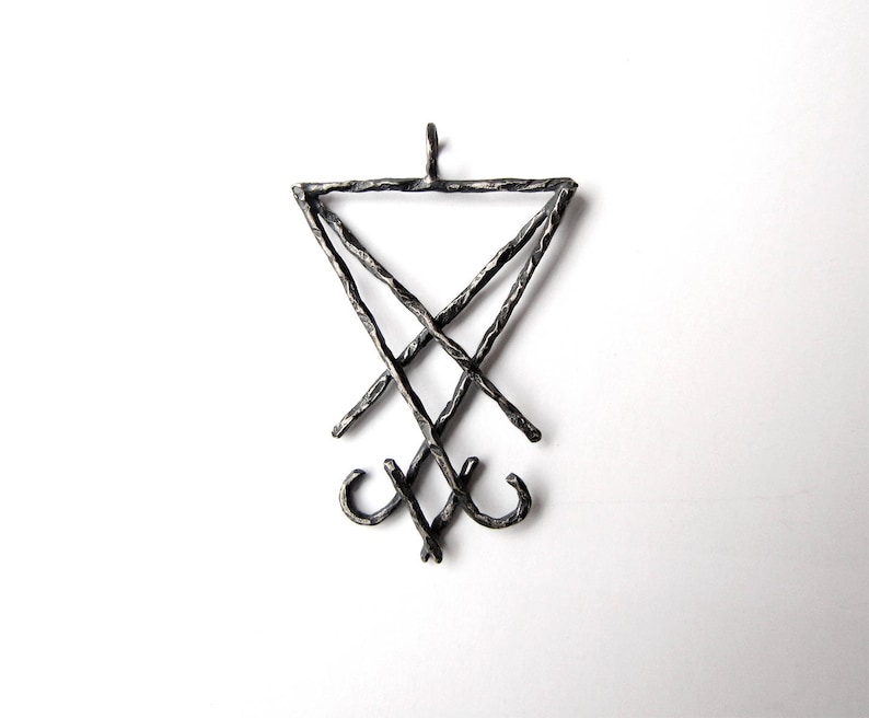 May include: A dark metal pendant featuring the Sigil of Lucifer symbol. The pendant has a triangular top with a cross and two curved lines at the bottom. The metal has a textured, handcrafted appearance.