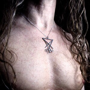 May include: A silver pendant necklace featuring a stylized, geometric design. The pendant hangs from a delicate chain, resting against a person's skin. The necklace is a dark, gothic style.