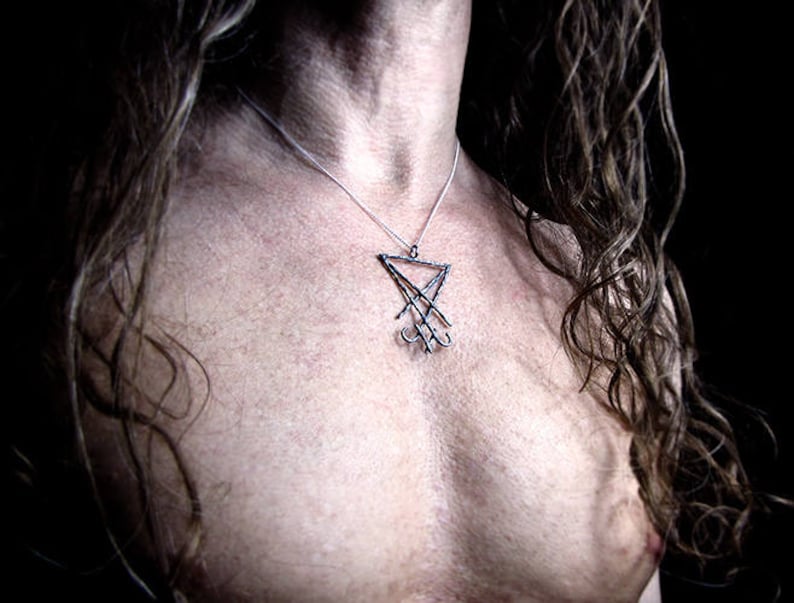 May include: A silver pendant necklace featuring a stylized, geometric design. The pendant hangs from a delicate chain, resting against a person's skin. The necklace is a dark, gothic style.