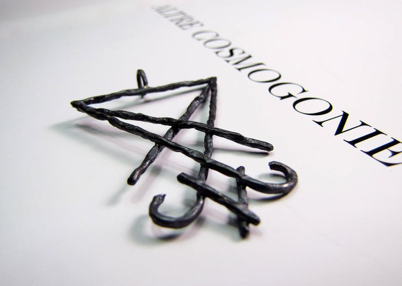 May include: A dark, textured metal pendant with an abstract design. The pendant has a loop at the top and curved elements. The word "AUTRE COSMOGONIE" is printed in black on the white background.