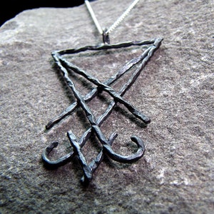 May include: A dark metal pendant necklace featuring the symbol of Leviathan. The pendant is a stylized, textured design with a triangular shape at the top and a cross-like element below. The necklace chain is silver.