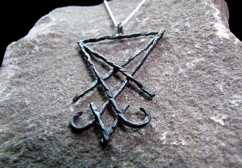 May include: A dark metal pendant necklace featuring the symbol of Leviathan. The pendant is a stylized, textured design with a triangular shape at the top and a cross-like element below. The necklace chain is silver.