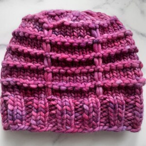 May include: A pink knitted hat with a textured pattern. The hat is made with a chunky yarn and has a slouchy fit.