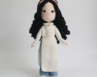 May include: A handmade crochet doll with long black curly hair, wearing a white headband with pink and green accents. The doll features a pale complexion, a white long-sleeved tunic with a high collar and a small pearl button, and blue trousers.