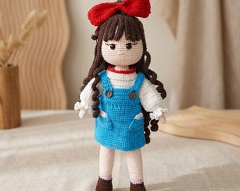 May include: A handmade crochet doll with long brown curly hair and a large red bow. The doll wears a white long-sleeved shirt with ruffled cuffs under a bright blue overall dress featuring two small brown buttons and two front pockets.