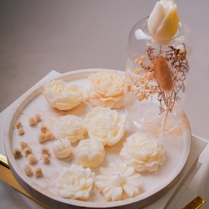Puede incluir: Velas con forma de flor de color marfil dispuestas sobre una bandeja redonda. Una campana de cristal exhibe una vela de tulipán y flores secas. Las velas, de varios tamaños y estilos, crean una presentación decorativa.