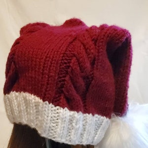 May include: A knitted burgundy hat with a white brim. The hat features a cable knit design and a soft, textured appearance. The white brim adds a contrasting element to the hat's overall design.