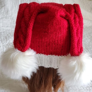 May include: Red knitted hat with white pom-poms and long, cable-knit ear flaps.