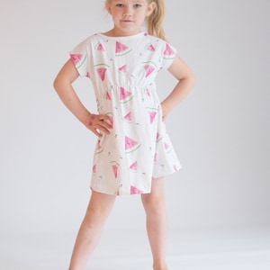 May include: A young girl wearing a white dress with a pink watermelon print. She is standing with her hands on her hips and looking at the camera.