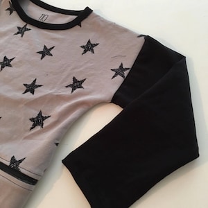Puede incluir: Una camiseta gris de manga larga con mangas negras y cuello negro. La camiseta presenta un estampado de estrellas negras y el número "110" impreso en el cuello.