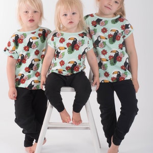 Könnte beinhalten: Drei Kinder tragen hellblaue T-Shirts mit einem Tukan- und tropischen Blumendruck. Die Shirts haben kurze Ärmel und einen Rundhalsausschnitt. Die Kinder tragen schwarze Hosen. Ein Kind sitzt auf einem weißen Stuhl.