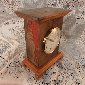 May include: A wooden clock with a gold face and black hands. The clock is made from reclaimed wood and has a rustic look. The clock is sitting on a white lace tablecloth.