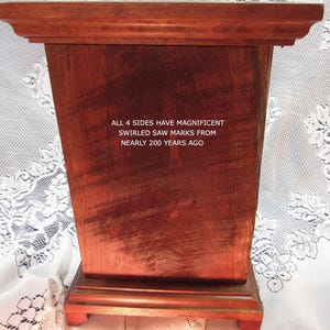May include: A rectangular wooden box with a reddish-brown finish. The box has a decorative top and sits on a smaller base. Text on the box reads: "ALL 4 SIDES HAVE MAGNIFICENT SWIRLED SAW MARKS FROM NEARLY 200 YEARS AGO."