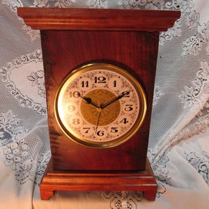 May include: A wooden mantel clock with a gold-colored clock face. The clock has a rectangular shape with a dark brown finish and a decorative base. The clock face features black numerals and hands.