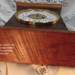 May include: A wooden clock with a round face and gold-colored trim. The clock face has black numerals and hands. The wooden base has visible swirled saw marks and the text "FANTASTIC 1830'S OLD SWIRLED SAW MARKS FROM ITS ORIGINAL SAW MILLING".