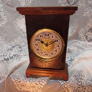 May include: A rectangular wooden clock with a round face. The clock face is white with black numerals and gold accents. The clock has a dark brown wooden frame and a lighter brown base.