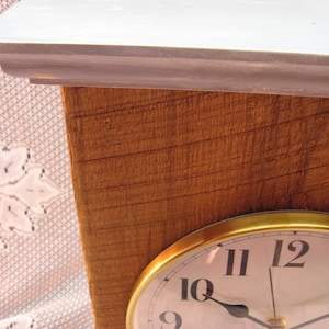 May include: A close-up of a wooden shelf with a gold-rimmed clock. The clock face is white with black numerals and hands. The shelf has a light gray top and a brown, textured wooden base.