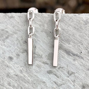 May include: A pair of silver earrings with a chain and a rectangular bar pendant.