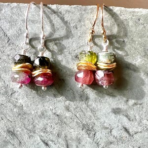 Może przedstawiać: Dwie pary kolczyków z kamieni szlachetnych. Jedna para ma srebrne haczyki, druga złote. Każdy kolczyk ma stos kolorowych, fasetowanych kamieni szlachetnych w odcieniach różu, zieleni i złota. Kolczyki są prezentowane na teksturowanej szarej powierzchni.