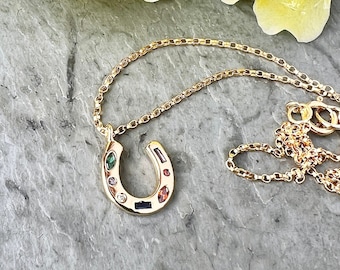 May include: A gold horseshoe necklace with colorful gemstones. The necklace is on a gray background with yellow flowers.