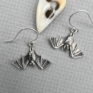 May include: A pair of silver bat earrings with detailed wings and hook closures. The earrings are displayed on a gray surface with a decorative, teardrop-shaped pendant in the background. The bat earrings are a unique accessory.