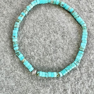 Könnte beinhalten: Türkis-Perlenarmband. Das Armband besteht aus kleinen, rechteckigen türkisfarbenen Perlen, die aufgefädelt sind. Die Perlen haben eine leuchtende türkisfarbene Farbe mit einigen Farbvariationen. Am Verschluss befindet sich eine silberfarbene Perle mit einem dekorativen Muster.