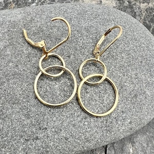 May include: A pair of gold-colored hoop earrings. Each earring features a lever back closure and three connected circular hoops of varying sizes. The earrings are displayed on a gray, textured surface.