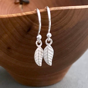May include: A pair of silver dangle earrings with small leaf charms. The earrings are hanging from a wooden bowl.