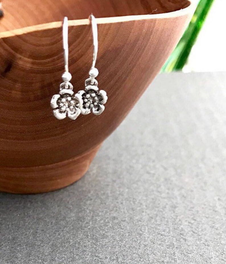 K&ouml;nnte beinhalten: Silberne Blumen-Ohrringe mit einem zarten Design. Die Ohrringe h&auml;ngen an einer Holzschale.