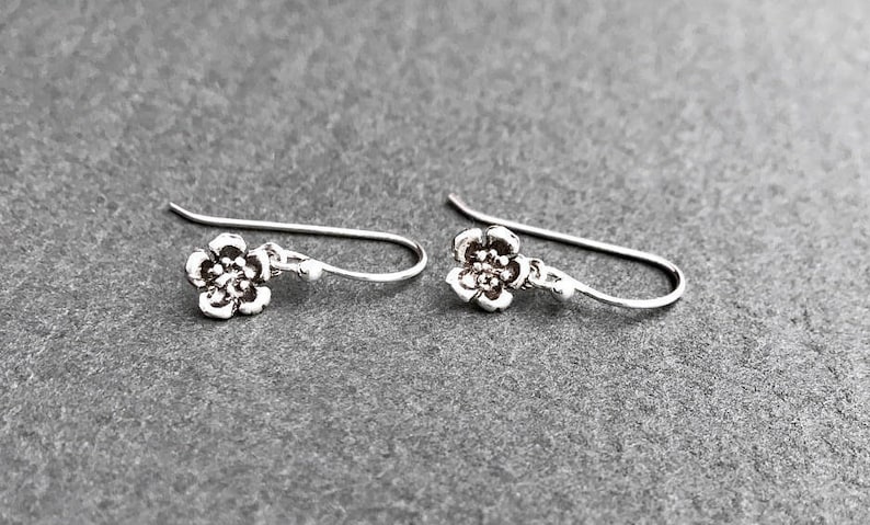 K&ouml;nnte beinhalten: Ein Paar silberne Ohrringe mit Blumen-Anh&auml;ngern. Die Ohrringe bestehen aus einem zarten Silberdraht und haben einen kleinen, detaillierten Blumenanh&auml;nger.