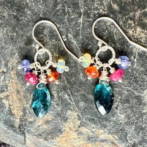 May include: A pair of handcrafted earrings with silver hooks. Each earring features a cluster of colorful gemstones, including blue, pink, orange, and iridescent beads, and a large, faceted teal teardrop-shaped stone.