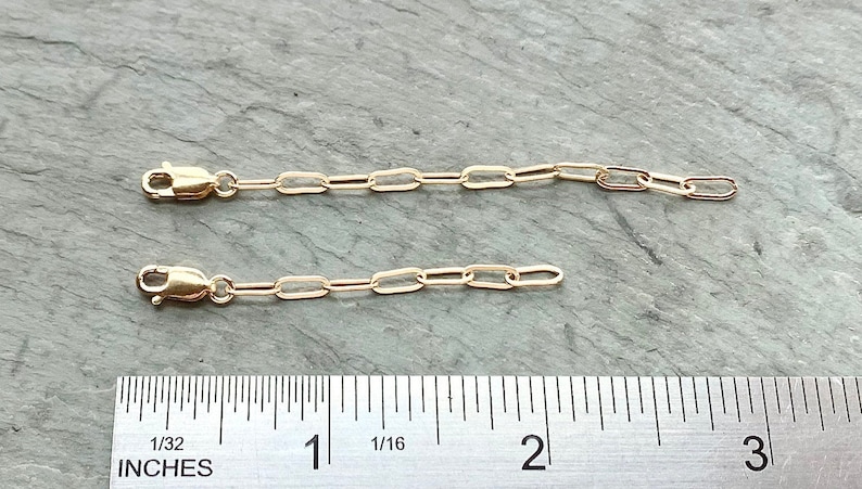 May include: Two gold chain extenders, each measuring 1 inch in length, with a lobster clasp on one end and a jump ring on the other. The extenders are shown next to a ruler.