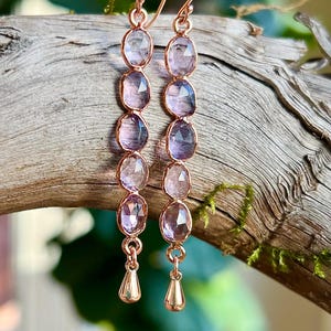 May include: Rose gold earrings with seven oval, faceted, light purple gemstones. The earrings have a teardrop-shaped charm at the bottom. The earrings are displayed on a piece of driftwood.