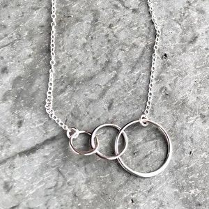 May include: A silver-toned necklace featuring a delicate chain and three interconnected circular rings of varying sizes. The rings are linked together in a horizontal row, creating a simple and clean design that hangs from the thin metallic chain.